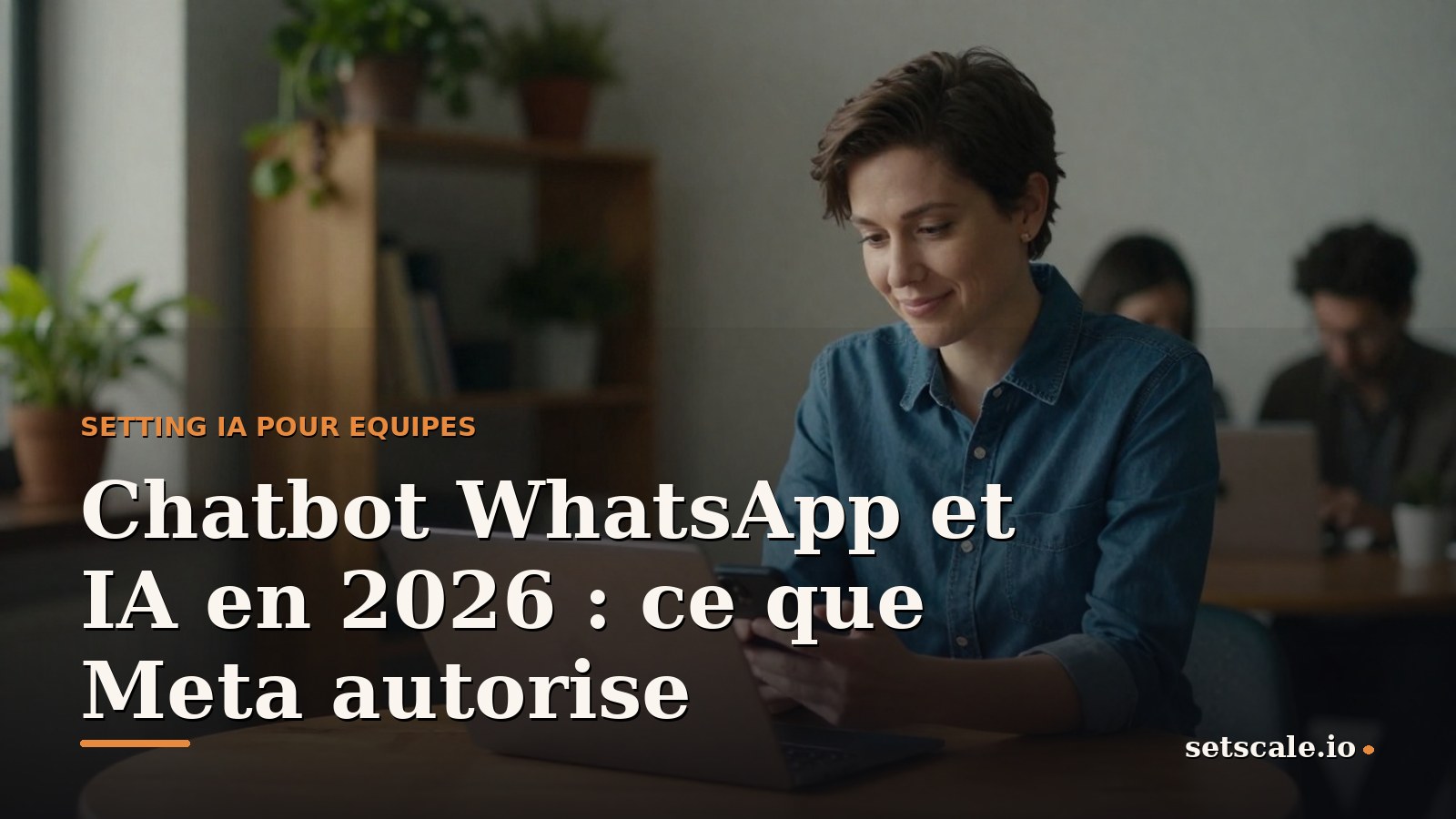 Homme d'une trentaine d'années aux cheveux noirs bouclés courts et à la barbe taillée, polo en maille bleu marine, assis à une table ronde en chêne dans un petit studio avec des plantes et des étagères en bois, téléphone en main, écran tourné vers lui, lumière naturelle chaude venant du côté